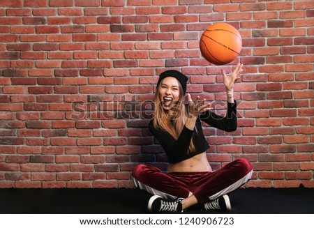 Photo of artistic girl 20s sitting on floor against brick wall and playing with basketball