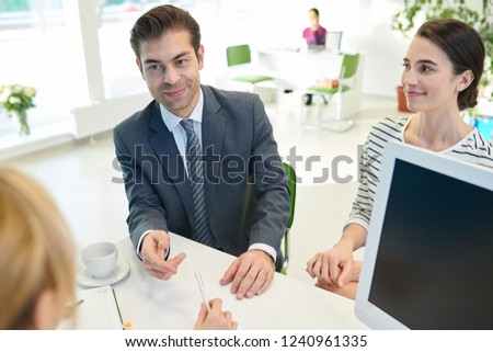 Smiling handsome businessman and his assistant sitting at table and talking to manager while signing investment contract with bank for business development
