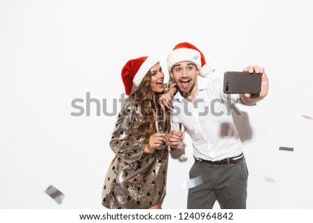 Cheerful young smartly dressed couple celebrating New Year party isolated over white background, taking a selfie, holding glasses of sparkling champagne