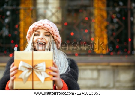 Laughing blonde model wearing fur coat and knitted cap pulls a gift box to you. Empty space
