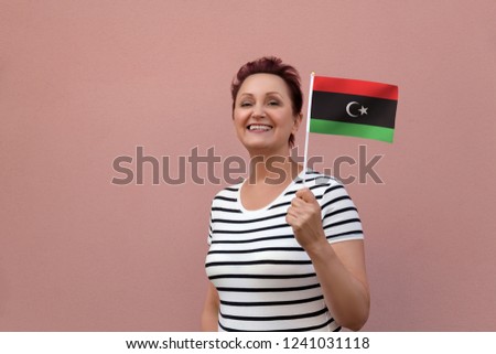 Libya flag. Woman holding Libyan flag. Nice portrait of middle aged lady 40 50 years old with a national flag over pink wall background outdoors.