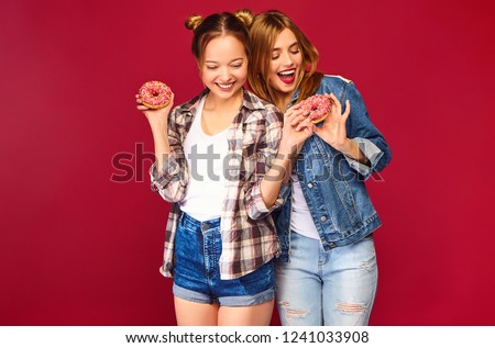 Two beautiful smiling hipster girls posing in trendy summer checkered shirt clothes.Women isolated on red background.Positive models holding fresh pink donuts with powder ready to enjoy sweets