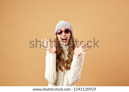 Girl dressed in white knitted sweater and hat and sunglasses is having fun on a beige background in the studio