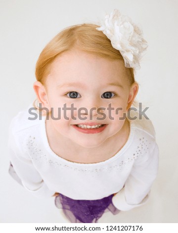 Portrait of a pretty little redheaded girl looking up at camera with a big smile, sparkly purple skirt, white shirt, and big flower hair clip. White background.