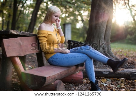 Young blonde woman sitting on a wooden bench and listening to music in the park.