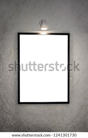 The picture in a black frame, hanging on the wall and illuminated by a lamp for paintings. On the background of a white wall, decorative structural plaster with glitter. Mock up.