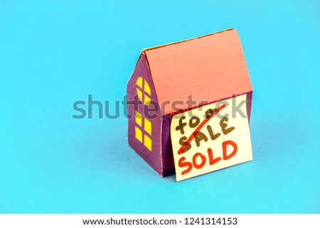 Purple cardboard house with yellow windows and sticker with word sold on blue background. Concept of real estate market