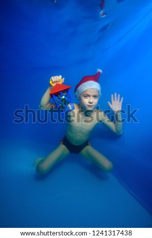 Portrait of a little boy who swims and poses underwater at the bottom of the pool in a red Santa hat with a toy in his hand on a blue background. Vertical orientation of the image