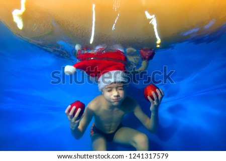 A little boy is swimming underwater with his eyes closed in a Santa Claus hat with red apples in his hands. Conceptual realism. Portrait. Landscape orientation of the image