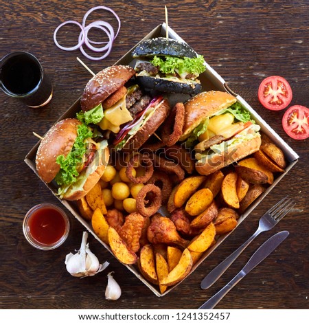 On a wooden background, 4 different burgers, various hot fried foods: potatoes, balls and deep-fried rings. around the glass with a drink, sliced tomatoes, cutlery, onion rings, red sauce and garlic. Royalty-Free Stock Photo #1241352457