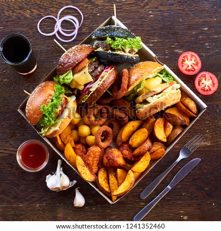 On a wooden background, 4 different burgers, various hot fried foods: potatoes, balls and deep-fried rings. around the glass with a drink, sliced tomatoes, cutlery, onion rings, red sauce and garlic. Royalty-Free Stock Photo #1241352460