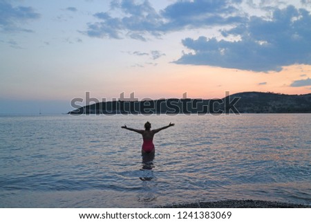Girl, seen from behind, in red swimsuit,  entering water to swim,  on the beach and watching beautiful sunset 