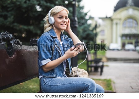 Young girl sitting on a bench in the city. Young beautiful woman listening to music with phone in outdoors.