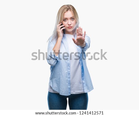 Young blonde woman talking using smarpthone over isolated background with open hand doing stop sign with serious and confident expression, defense gesture