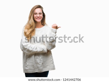 Young beautiful blonde woman wearing winter sweater and sunglasses over isolated background smiling with happy face looking and pointing to the side with thumb up.