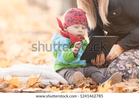 Mother and baby boy sitting on a blanket in the park using digital tablet. Young mom teach his child how to use tablet with touchscreen. Small child quickly learns to handle new technological things.