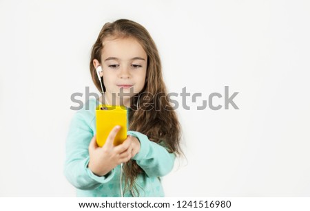 Bright beautiful little girl in headphones with the phone emotionally posing for a selfie on an isolated background.