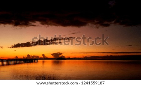 Orange and yellow hues sunset with low dark clouds over the water with reflection, background with a fishing pier jetty, serene tranquil backdrop, seascape cloudscape, natural view