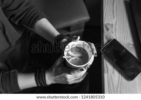 Top view. Student woman holds with two hands a hot cup of tea with sea buckthorn Hippophae and a slice of orange. Workplace, notebook with pen and laptop, mobile phone, smartphone, black and white