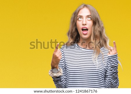Beautiful young blonde woman wearing stripes sweater over isolated background amazed and surprised looking up and pointing with fingers and raised arms.