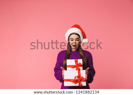 Image of a shocked young emotional woman posing isolated over pink background holding gift boxes wearing christmas hat.