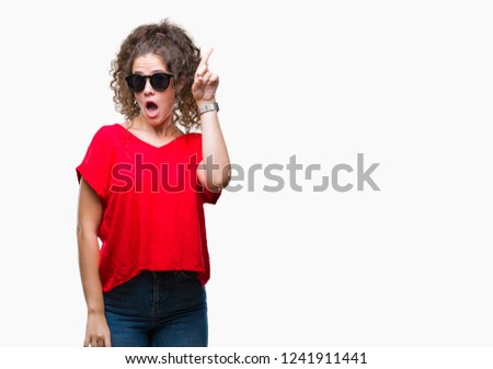 Beautiful brunette curly hair young girl wearing sunglasses over isolated background pointing finger up with successful idea. Exited and happy. Number one.