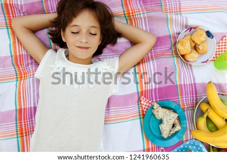 Overhead portrait of beautiful child girl laying on blanket having picnic food, relaxing smiling with eyes closed, outdoors. Fruit and sandwiches fun healthy eating, leisure recreation lifestyle.