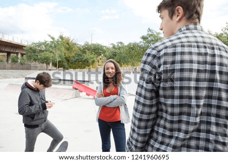 Group of diverse beautiful trendy teenagers in sports ground skating park with skateboard relaxing using technology, outdoors. Smiling students in grunge urban lifestyle, recreation leisure.
