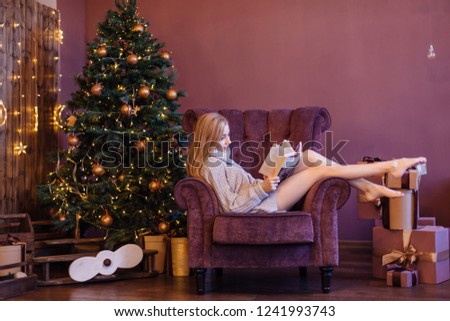 Beautiful young woman dressed in a sweater sitting on the cozy arm chair next to the christmas tree with gifts and reading book. Christmas cozy photo