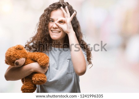 Young brunette girl holding teddy bear over isolated background with happy face smiling doing ok sign with hand on eye looking through fingers