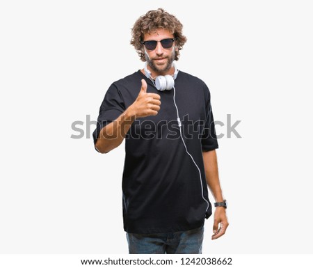 Handsome hispanic man listening to music wearing headphones over isolated background doing happy thumbs up gesture with hand. Approving expression looking at the camera with showing success.