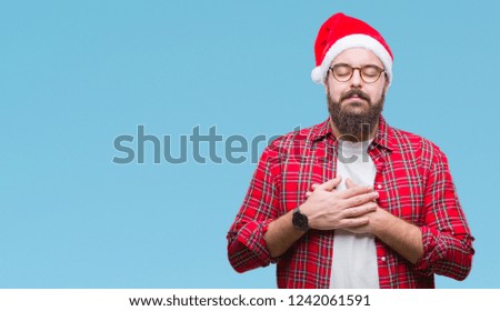 Young caucasian man wearing christmas hat over isolated background smiling with hands on chest with closed eyes and grateful gesture on face. Health concept.