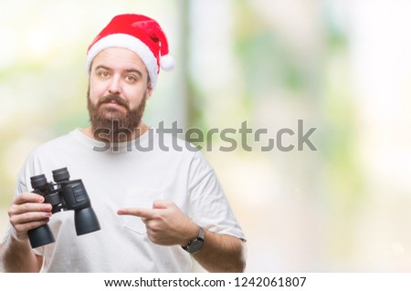 Young caucasian hipster man wearing christmas hat looking though binoculars over isolated background very happy pointing with hand and finger