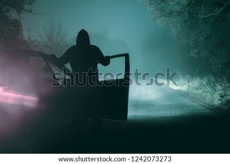 A lone, hooded figure standing next to a car looking at an empty misty winter country road silhouetted at night by car headlights. With a cold, grainy, muted edit.