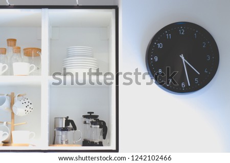 Black wall clock, white glass plate and coffee maker in the kitchen. Do not focus on objects.