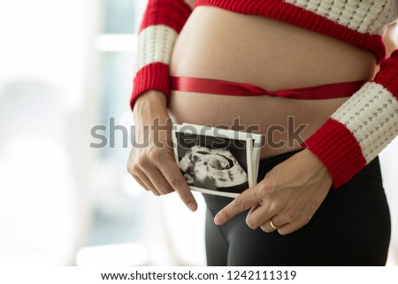 Close up of pregnant woman holding ultrasound scan image film on her tummy with red Ribbon on belly for love and happiness,Healthy Pregnant Concept,Selective Focus