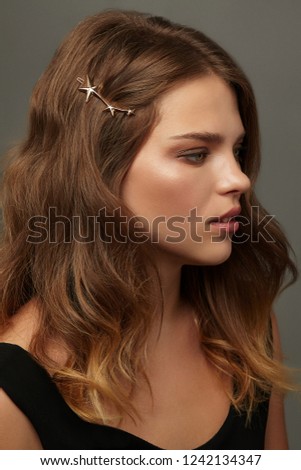 Close up portrait of a young brunette lady with natural make-up and wavy hairstyle. Her tresses are adorned with golden stars hair clip. The girl posing on the grey background, looking to the side. Royalty-Free Stock Photo #1242134347
