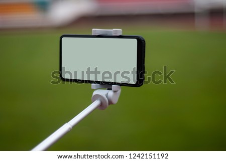 Mock up of a black smartphone with a selfie stick. White screen for the design of the template on the background of a green lawn on the football field