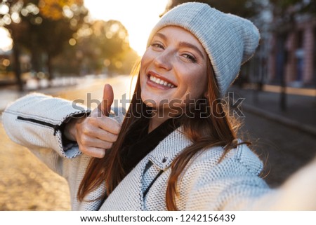 Beautiful young woman dressed in autumn coat and hat walking outdoors, taking a selfie