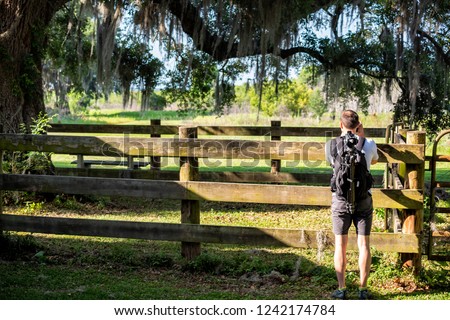 Back of young man, nature photographer standing, taking pictures of landscape in Gainesville, USA Paynes Prairie Preserve State Park in Florida, wooden fence paddock stable