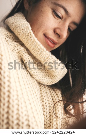 Stylish hipster girl in knitted sweater smiling in soft light in room. Portrait of young attractive woman relaxing and taking selfie. Atmospheric cozy winter moments. Moody image