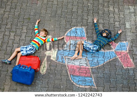 Two little children, kids boys having fun with with airplane picture drawing with colorful chalks on asphalt. Friends painting with chalk and going on vacations dreaming of pilot profession.