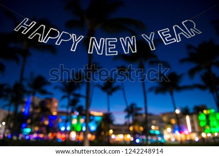 Happy New Year message hanging in glittery party bunting above tropical palm trees of the glowing neon lights of the skyline of Miami Beach, Florida, USA