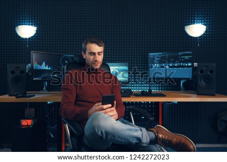 Young man working in the studio using a smartphone and computers. Caucasian freelancer holding mobile phone sitting leg over leg.