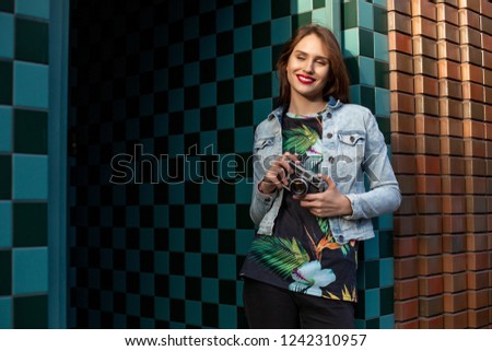 Cool funny girl model with retro film camera wearing a denim jacket, dark hair outdoors over city wall in a cage background