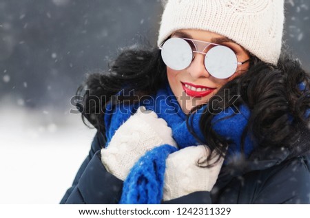 Outdoors lifestyle close up portrait of beautiful girl walking in the snowy winter park. Smiling and enjoying wintertime. Wearing stylish mirrored sunglasses, down jacket, knitted hat. It's snowing