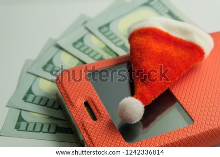 Souvenir Santa Claus hat lying on a smartphone in a red case against the background of five hundred US dollars. Close-up. The concept of New Year's recharge mobile phone or a gift for Christmas.