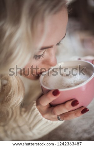 Cozy home portrait of a young beautiful woman. A beautiful blonde girl in a knitted dress is sitting by the window drinking a cappuccino and reading a book. Autumn decor. Cozy. Winter. Autumn.