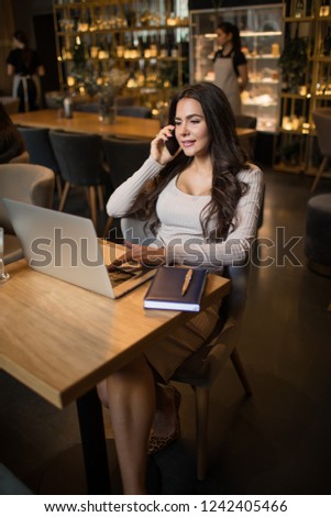 Happy smiling woman entrepreneur having pleasant cell telephone conversation during work on laptop computer while sitting in restaurant during work break. Female broker talking with client 