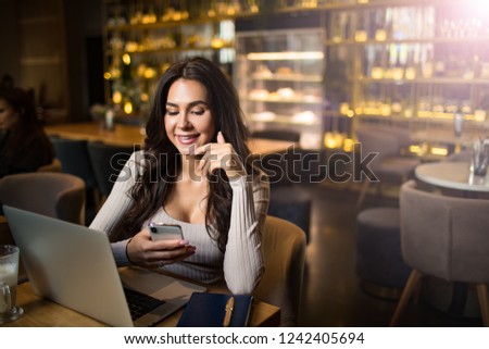Beautiful smiling charming woman associate web editor using apps on mobile phone during distance work on laptop computer while sitting in restaurant near copy space for promotional content. 